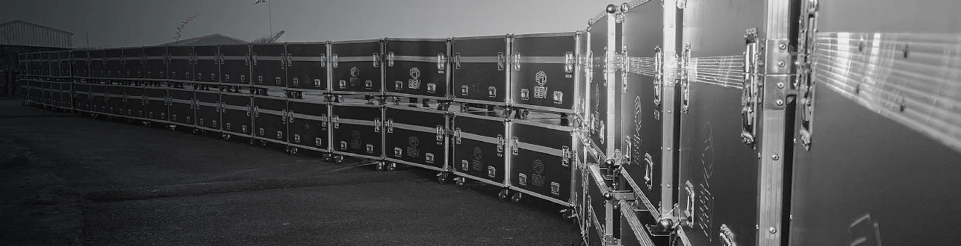 A long row of large black road cases with metal edges and wheels are lined up outdoors on a paved surface.