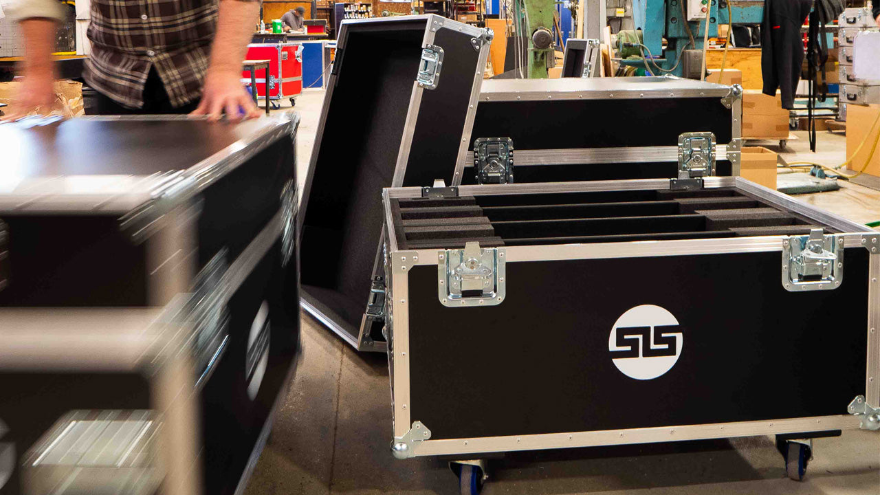A person moves a large black equipment case near other similar cases with metal edges and handles in an industrial workshop.