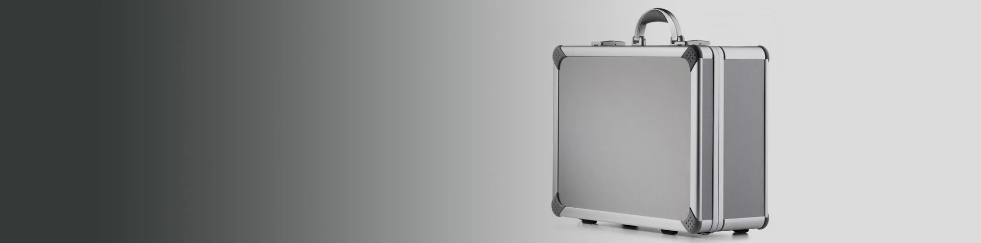 A silver metal briefcase with reinforced corners stands upright against a light gray background, with a gradient fading to dark gray on the left.