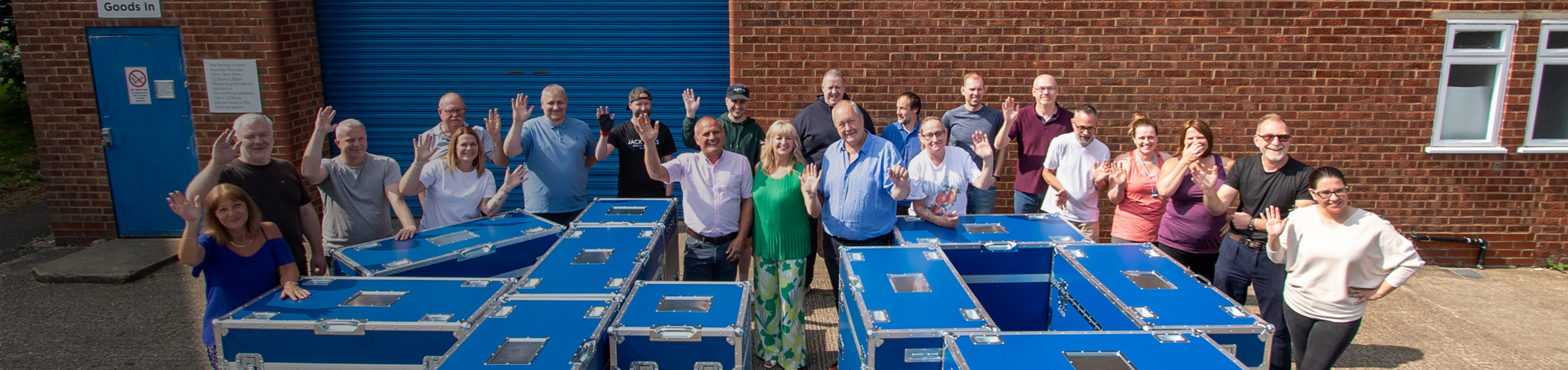 A group of people stand outdoors next to several large blue containers, waving at the camera in front of a brick building with a blue roller door.