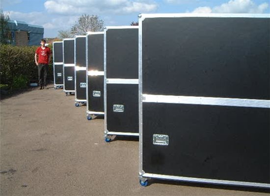 A row of large black road cases with silver trim on wheels is lined up outdoors on a paved surface; a person stands in the background.