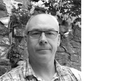 A middle-aged man with glasses and a plaid shirt stands in front of a stone wall with flowers in black and white.