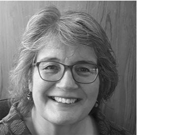 A woman with short, gray hair and glasses smiles at the camera. She is wearing earrings and a patterned top, and the background is a wooden surface.