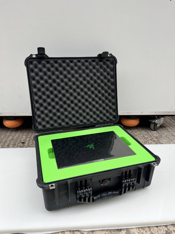 A black hard case with foam padding holds a closed black laptop embedded in bright green foam, placed on a white surface outdoors.