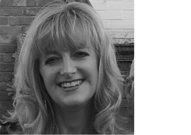 A woman with shoulder-length wavy hair smiling, posed in front of a brick and tile wall. The image is in black and white.