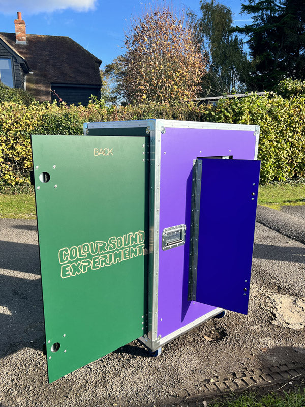 A large box labeled COLOUR SOUND EXPERIMENT with green and purple doors stands open on a paved outdoor area near grass and trees.