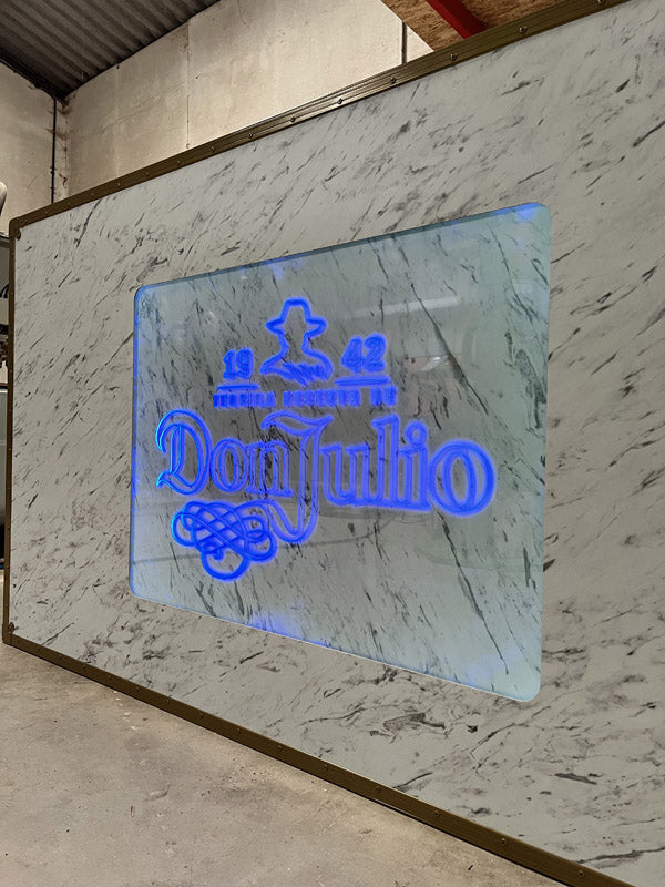 A marble wall with a glass panel displays a blue illuminated Don Julio 1942 logo and emblem.