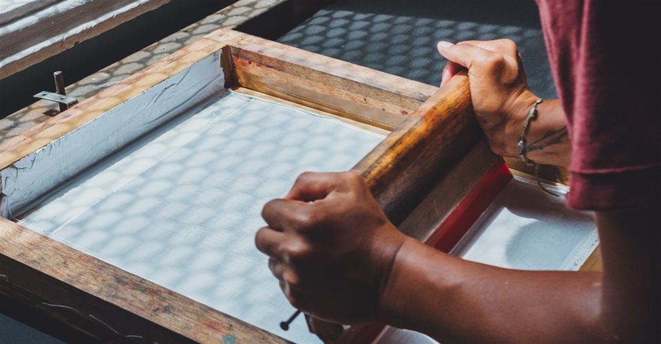 Person using a wooden frame and squeegee for screen printing on a flat white surface under natural light.