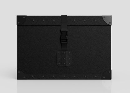 The Standard Cube Storage Box - Small is a black, rectangular trunk featuring metal corner protectors, a central latch, visible edge rivets, and a strong webbing buckle, shown against a plain light background.