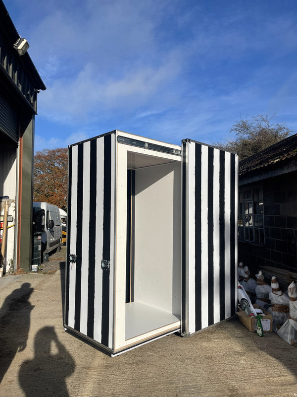A black and white striped booth with open doors stands outside on concrete near industrial buildings, with bags and various items nearby.